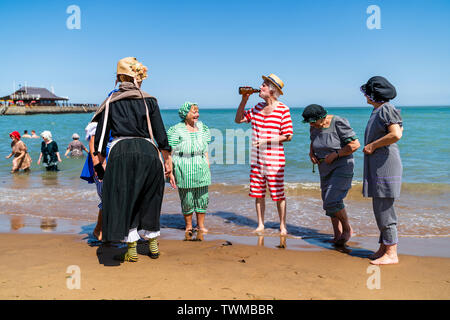 Broadstairs Dickens Festival. Cinq hauts hommes et femmes vêtus de costumes de bains Victorienne, debout sur le bord de la mer à la plage de la baie de Viking. Homme debout au milieu de boire une bière d'une bouteille, d'autres l'observant. Banque D'Images