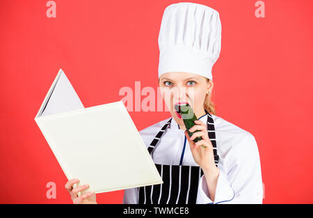 Woman in hat cook et tablier. chef professionnel dans la cuisine manger du concombre. Cuisine. happy woman la cuisson des aliments sains par recette. Femme au foyer avec livre de cuisine. menu du restaurant. Suivre un régime culinaire. Banque D'Images