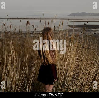 Le retour d'une femme avec de longs cheveux blonds promenades à travers les hautes herbes vers le Grand Lac Salé au coucher du soleil. Banque D'Images