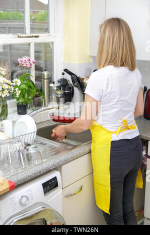 Une jeune femme en tablier jaune lave la vaisselle et nettoie la cuisine. La femme de ménage lave le pate dans l'évier. Vie domestique Banque D'Images