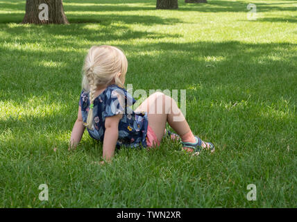 Petite fille en rose short et t-shirt bleu à motifs assis dans l'herbe verte avec les genoux fléchis. Son dos est à l'appareil photo. Banque D'Images