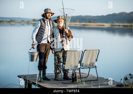 Avec grand-père fils adultes debout ensemble sur la jetée en bois, profiter du lever du soleil alors que la pêche sur le lac tôt le matin Banque D'Images