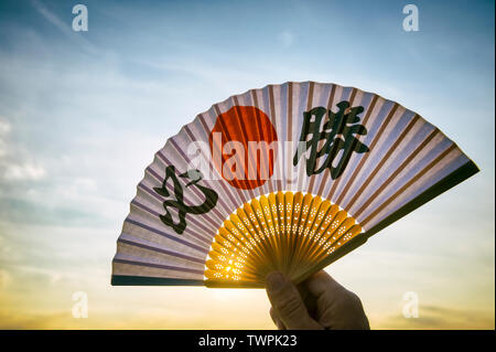 De la main tenant un supporteur sport japonais fan décoré de caractères kanji énonçant hissho (traduction anglaise : certaines victoire) au lever du soleil Banque D'Images