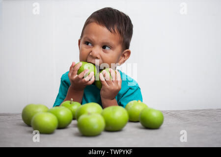 Un petit garçon ayant une collation saine par ardemment en essayant d'ajuster et de mordre deux pommes vertes à la fois dans sa bouche, avec un tas de pommes vertes à l'avant o Banque D'Images
