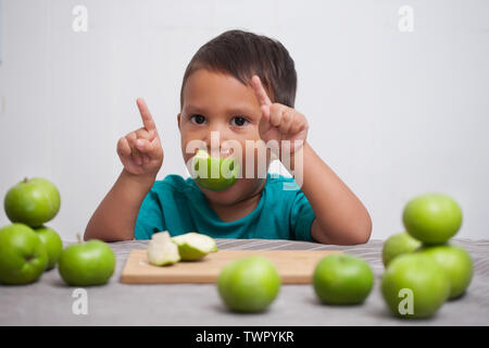 Un mignon jeune garçon signalant avec ses mains qu'il aime bien manger les fruits dans son alimentation, et tenant une pomme verte dans sa bouche. Banque D'Images