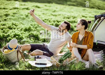 Les jeunes et joyeux couple photo selfies lors du pique-nique à le camping, les voyages en montagne au coucher du soleil Banque D'Images