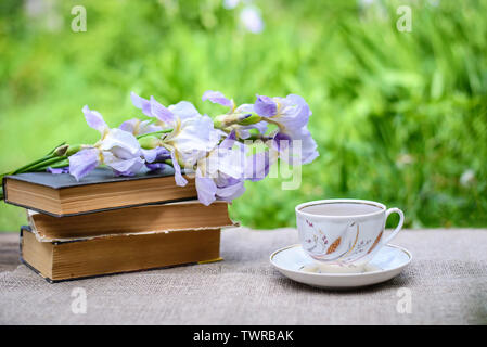 Une pile de vieux livres, fleurs pourpres iris et une tasse de thé sur un feuillage vert d'été contexte Banque D'Images