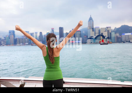 Happy woman cheering succès par Hong Kong skyline with arms raised up tendus. Gagnant la célébration joyeuse sur la promenade de Tsim Sha Tsui et de l'Avenue des Stars à Victoria Harbour, Kowloon. Banque D'Images