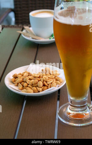 Un verre de bière de lumière froide, d'arachides dans une soucoupe blanche et une tasse de americano sur une table en bois. Banque D'Images