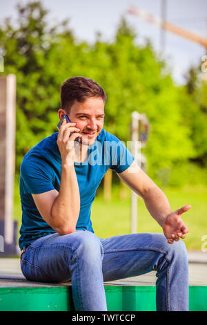 Beau jeune homme est assis à l'extérieur et de parler avec quelqu'un au téléphone Banque D'Images