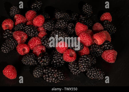 Framboises mûres fraîches et de petits fruits noirs sur fond noir close up avec de l'eau gouttes Banque D'Images