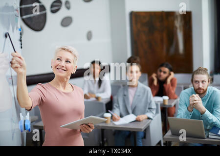 Waist up portrait of smiling female teacher pointing at whiteboard lors de la présentation de cours de formation pour étudiants en classe, copy space Banque D'Images
