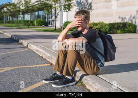 Adolescent bouleversé reposant sa tête sur sa main, comme il est assis sur le trottoir en face de son école. Banque D'Images