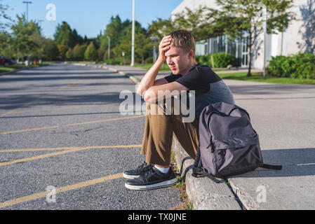 Adolescent bouleversé reposant sa tête sur sa main, comme il est assis sur le trottoir en face de son école. Banque D'Images