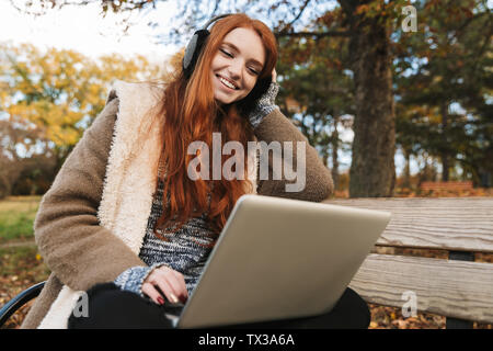Belle jeune fille rousse de l'écoute de musique avec headpones, assis sur un banc, à l'aide d'un ordinateur portable Banque D'Images