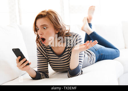 Jolie jeune femme choquée à l'aide de téléphone mobile tout en portant sur un canapé Banque D'Images