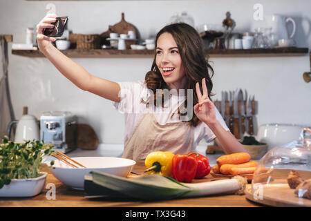 Belle jeune femme portant un tablier cuisine salade saine à la cuisine à la maison, en tenant un téléphone mobile avec des selfies Banque D'Images