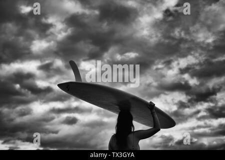 Femme surfer carrying board long passage sous ciel nuageux Banque D'Images