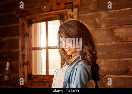 Jeune femme avec de longs cheveux smiling près de la fenêtre dans une maison de campagne. Banque D'Images