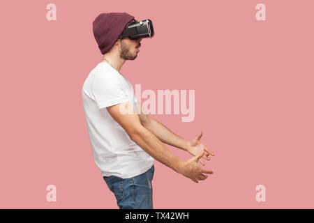 Vue de côté portrait de jeune homme barbu choqué en chemise blanche, casual hat standing, portant des vr, jouant le jeu vidéo et essayer de toucher quelque chose. Ind Banque D'Images