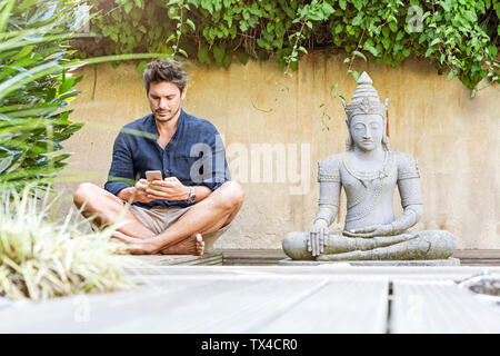 Homme assis en tailleur à côté de la statue de Bouddha dans un jardin Zen, using smartphone Banque D'Images