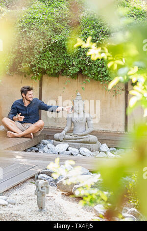 Homme assis en tailleur à côté de la statue de Bouddha dans un jardin Zen, using smartphone Banque D'Images