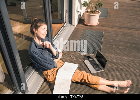 Jeune femme aux yeux clos assis sur la terrasse à la maison avec un ordinateur portable et réservez Banque D'Images