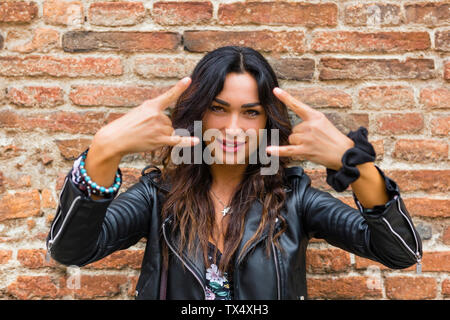 Portrait de jeune femme portant veste en cuir noire et montrant des signes de rock and roll, mur de brique Banque D'Images