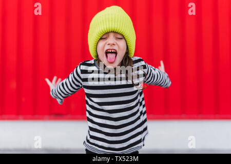 Portrait of little girl shirt et capuchon jaune sticking out tongue en face de fond rouge Banque D'Images