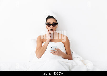 Image d'une belle surprise avec femme émotionnelle towel on head se trouve au lit sous couverture isolated over white wall background portant des lunettes de cha Banque D'Images