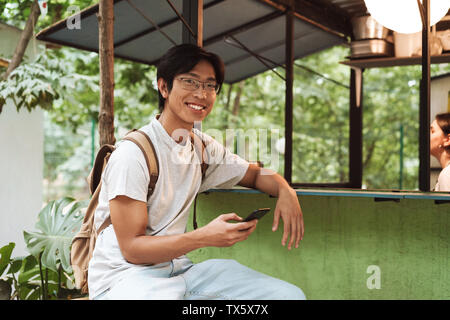 Smiling asian student homme portant un sac à dos à l'extérieur, assis, à l'aide de mobile phone Banque D'Images