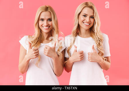 Smiling blonde jumeaux portant en t-shirts showing Thumbs up et regardant la caméra sur fond rose Banque D'Images