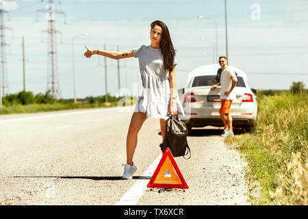 Le jeune couple a enfoncé la voiture tout en voyageant sur la façon de se reposer. Ils tentent d'arrêter les autres conducteurs et demander de l'aide ou l'auto-stop. Relation, problèmes sur la route, en vacances. Banque D'Images