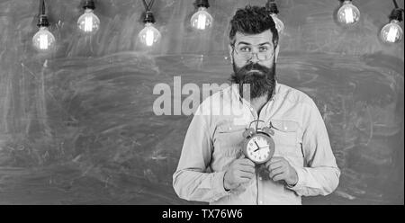 Calendrier scolaire concept. Homme avec barbe et moustache sur le visage calme de stand de classe. Hipster barbu est titulaire d'réveil, tableau sur toile de fond, l'espace de copie. Enseignant dans les lunettes est titulaire d'un réveil. Banque D'Images