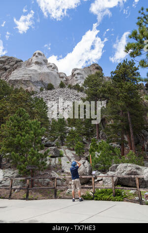 Un jeune homme se distingue par une balustrade à la base de la montagne pour prendre une photo du mont Rushmore de pins et de ciel bleu. Banque D'Images