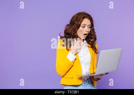 Portrait d'une jolie jeune femme rousse choqué isolés sur fond violet, using laptop Banque D'Images