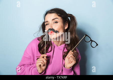 Portrait de la princesse brunette girl holding paper fake moustache et lunettes de stick isolé sur fond bleu en studio Banque D'Images