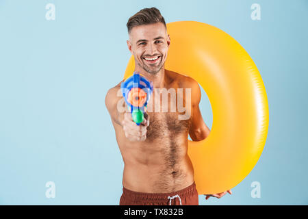 Photo d'un beau émotionnel excité hot man isolated over blue wall background avec anneau en caoutchouc beach holding water gun jouet. Banque D'Images