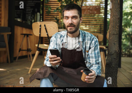 Photo de jeune serveur man wearing apron using smartphone tout en travaillant dans un café ou café, piscine Banque D'Images
