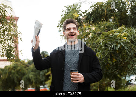 Photo de l'homme gai 30s veste port holding newspaper et café à emporter tout en marchant à travers green park Banque D'Images