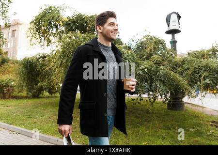 Photo de jeune homme 30s veste port holding newspaper et café à emporter tout en marchant à travers le parc de la ville Banque D'Images