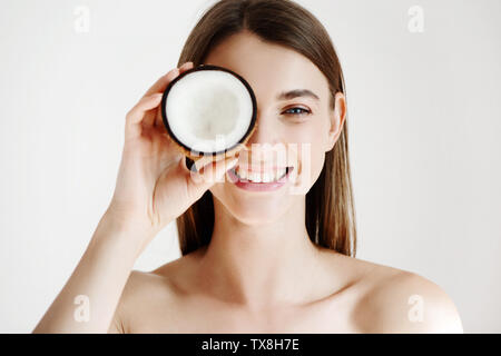 Portrait of beautiful smiling woman with nettoyer la peau parfaite de se cacher derrière des yeux la moitié de noix de coco sur fond blanc. Banque D'Images
