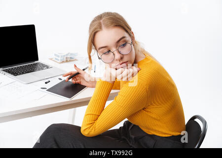 Vue latérale d'une belle jeune femme fatiguée graphic designer assis sur le lieu de travail travaillant sur écran blanc ordinateur portable et tablette sur isolés Banque D'Images
