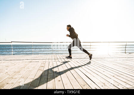 Photo de jeunes sportifs en survêtement noir 20s'exécute en station le long trottoir de bois au cours d'exercices du matin Banque D'Images