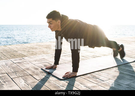 Photo de concentrés sportsman 20s en survêtement femme couchée sur le tapis de fitness et faire de l'exercice de planches tout en travaillant par mer dans la matinée Banque D'Images