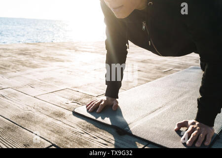 Photo de strong sportsman 20s en survêtement femme couchée sur le tapis de fitness et faire de l'exercice de planches tout en travaillant par mer dans la matinée Banque D'Images