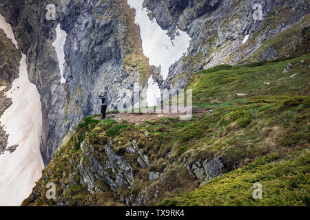 Le randonneur, la protection d'un vent avec une capuche, debout sur le bord d'une falaise sur la montagne de Rila en Bulgarie et de photographier avec son téléphone Banque D'Images