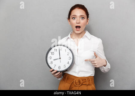 Image de femme rousse émotionnelle 20s dans l'usure formelle demande à huis clos en maintenant grosse horloge sur fond gris isolé Banque D'Images