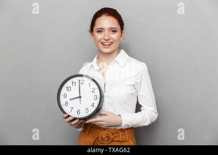 Image de belle femme rousse 20s dans l'usure formelle demande à huis clos en maintenant grosse horloge sur fond gris isolé Banque D'Images