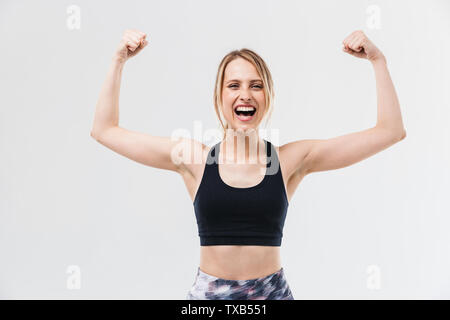 Image de femme blonde excité 20s'habiller dans les vêtements de sport de l'exercice et de faire les exercices avec haltères courtes au cours de fitness dans une salle de sport isolated over white wall Banque D'Images